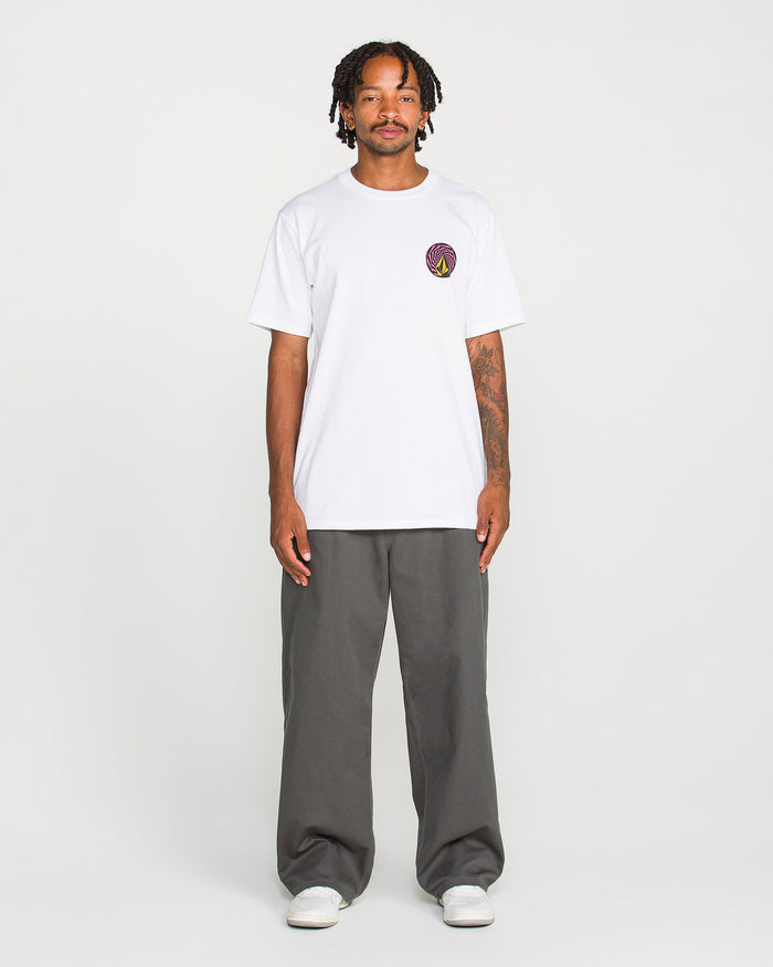 A person stands facing forward, wearing the Volcom Men's Dirty Work Short Sleeve Tee—a classic fit made from heavyweight cotton with a small soft-hand screen print logo on the left chest—paired with gray pants and white shoes, against a plain white background.