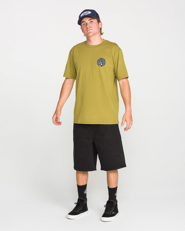 Wearing a classic fit green Men's Dirty Work Short Sleeve Tee by Volcom, paired with black knee-length shorts, black socks, black sneakers, and a dark blue cap, a person stands against a plain white background.