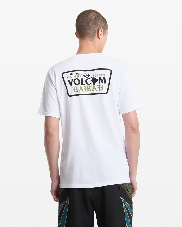 A man with short hair stands with his back to the camera, wearing Volcom’s Men's Dispatch Short Sleeve Tee in white, featuring a Hawaii license plate graphic on the back, paired with dark patterned shorts.