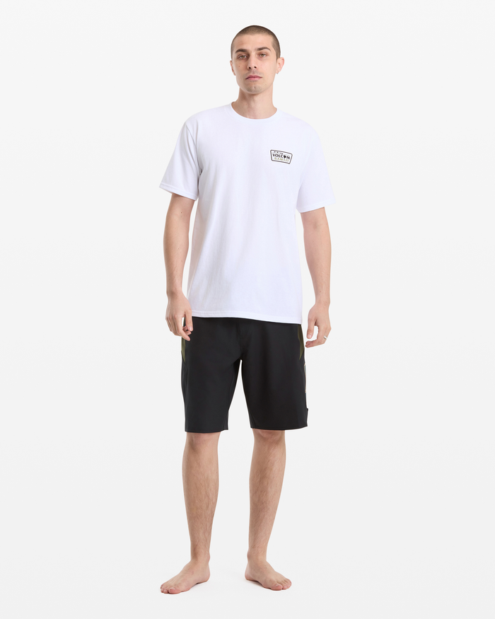 A man stands barefoot against a white background, wearing the Volcom Men's Dispatch Short Sleeve Tee in white and black knee-length shorts. He has short hair, a neutral expression, and his arms are relaxed at his sides.
