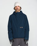 A person with a mustache and curly hair wears a white beanie, gray pants, patterned gloves, and the Volcom Men’s Double Insulated Jacket featuring Zip Tech in navy blue, standing against a plain white background.