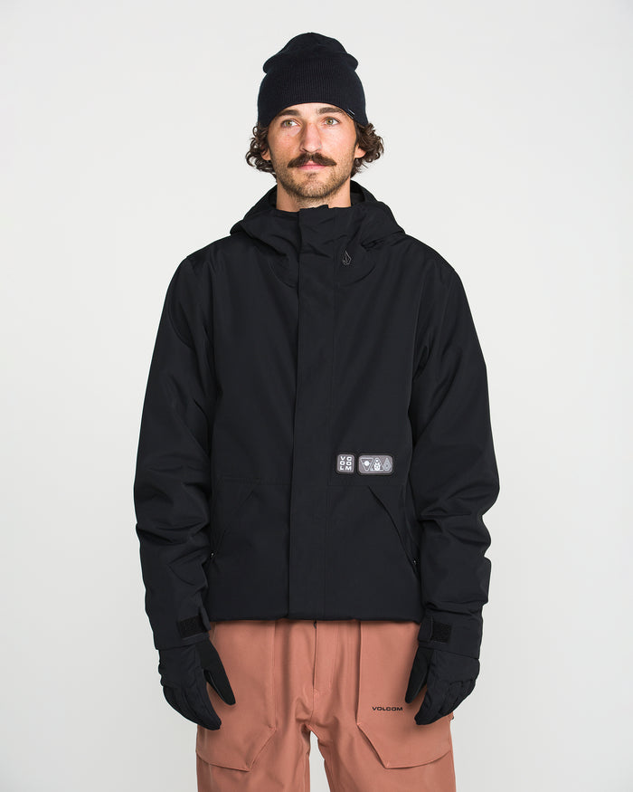 A man with a mustache, wearing a black beanie, black Volcom Men’s Double Insulated Jacket, black gloves, and light brown pants stands facing the camera against a plain white background.