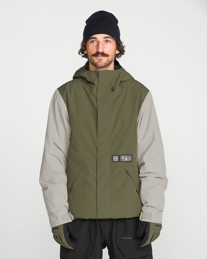 A man with a mustache wears a black beanie, black gloves, and the Volcom Men’s Double Insulated Jacket—an olive green and gray technical outerwear piece featuring recycled warmth—as he stands facing forward against a plain white background.