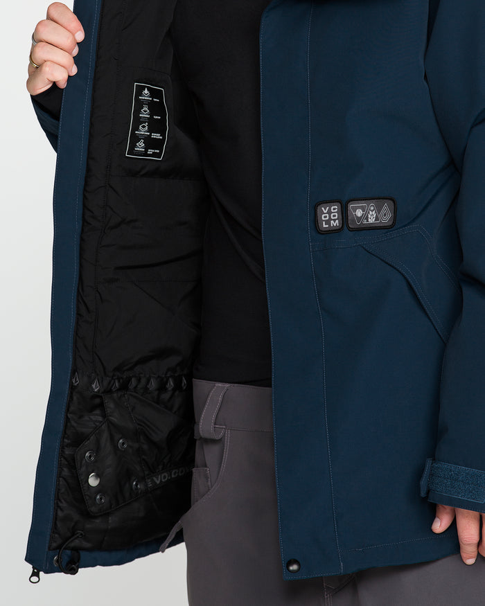 A man wearing gray pants and a dark shirt opens the Volcom Men’s Double Insulated Jacket in blue, exposing its black inner lining detailed with labels, pockets, and innovative features like Zip Tech. The image focuses on his lower face and torso, emphasizing the jacket’s recycled warmth construction.