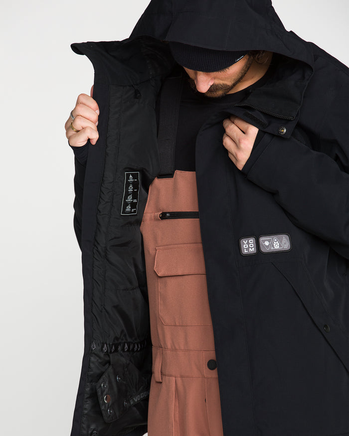 A person looks down while opening a Volcom Men’s Double Insulated Jacket in black, revealing the inner labels. They are wearing a black shirt and brown overalls against a plain white background.