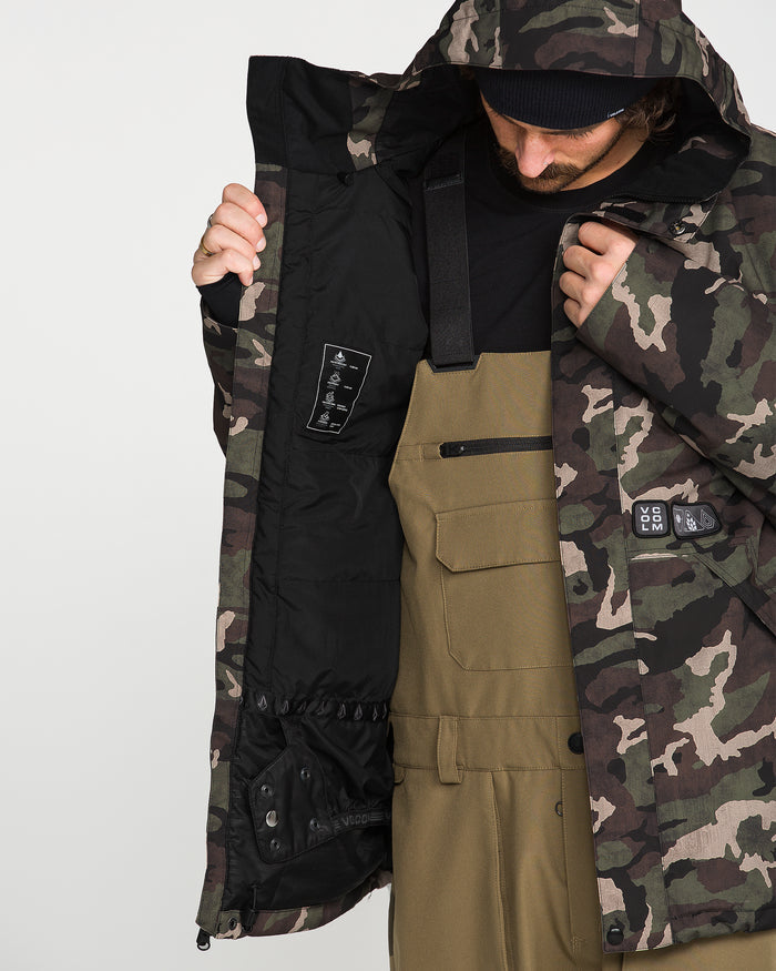 Wearing the Volcom Men’s Double Insulated Jacket, a person in brown overalls opens the camouflage technical outerwear to show its black inner lining and a label featuring symbols that emphasize its 10K/10K V-Science performance. With their face partly concealed by the jacket’s hood and a beanie, they highlight both recycled warmth and functional design.