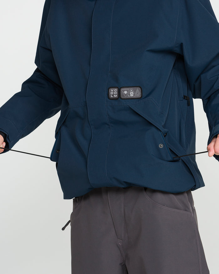 A person is shown wearing the Volcom Men’s Double Insulated Jacket in dark blue paired with dark pants, adjusting the elastic drawstring at the hem of the jacket. The image focuses on the torso and arms; the head is not visible.