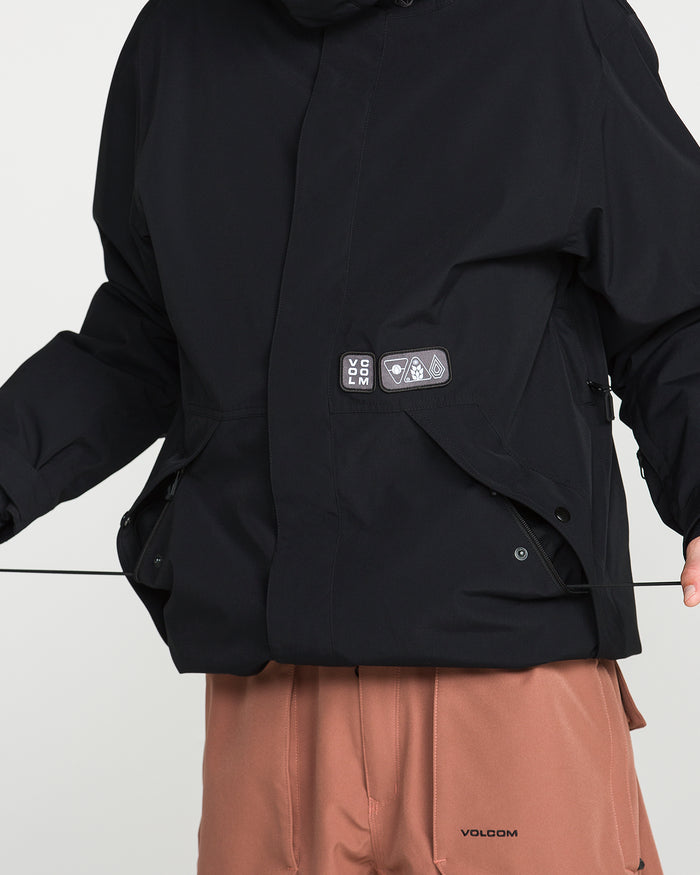 A person wearing a black Volcom Men’s Double Insulated Jacket with patch details and light brown pants featuring a small Volcom logo holds a black string with both hands. The person’s face is not visible.