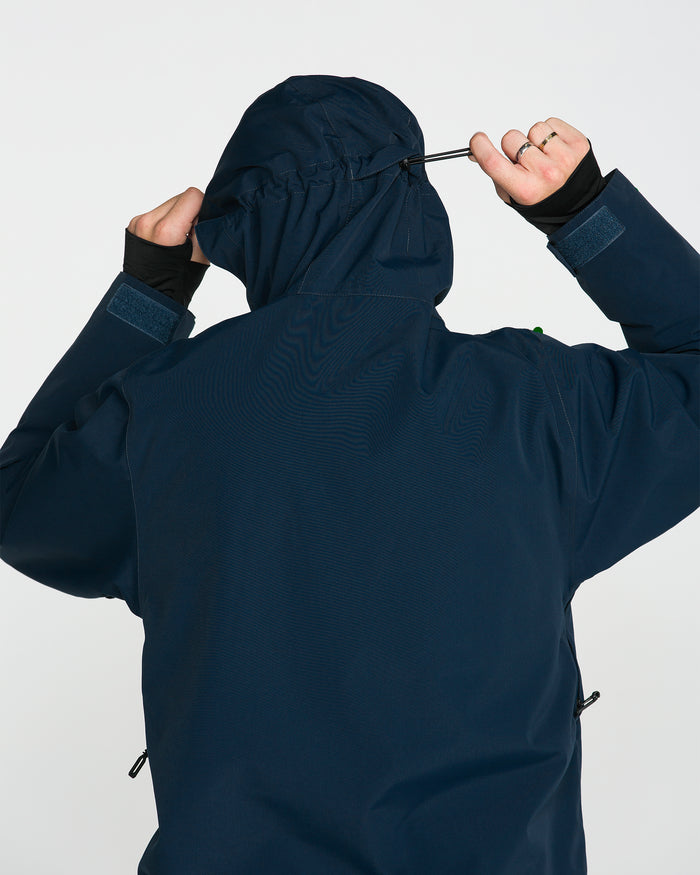 A person in the Volcom Men’s Double Insulated Jacket, shown from behind, tightens the hood’s drawstring. The plain, light background highlights the jacket’s recycled warmth and modern technical design.