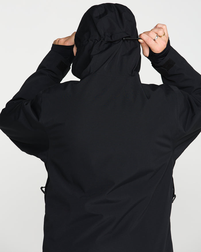 A person seen from behind adjusts the hood of the Volcom Men’s Double Insulated Jacket, a black technical outerwear piece, against a plain light background.