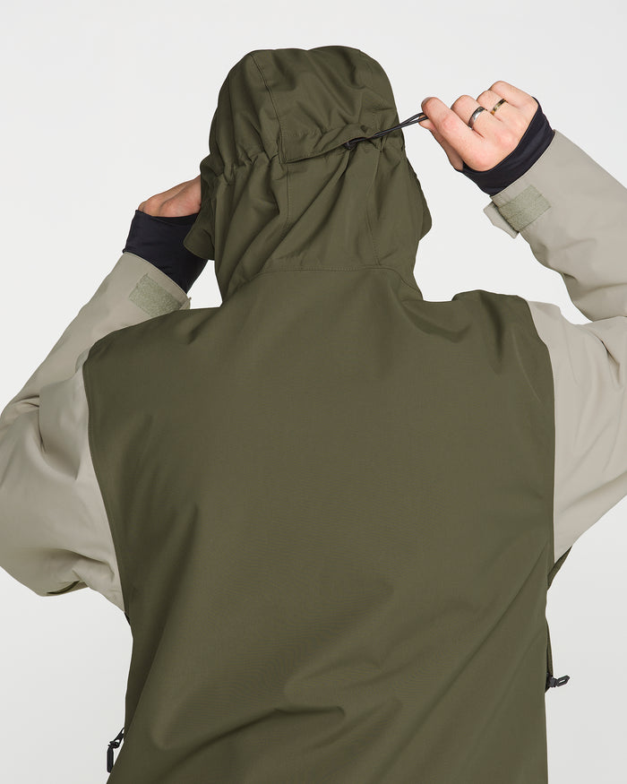 A person is seen from behind adjusting the hood of the Volcom Men’s Double Insulated Jacket in olive and beige, pulling a drawstring with one hand while wearing a gold ring, against a plain white background.
