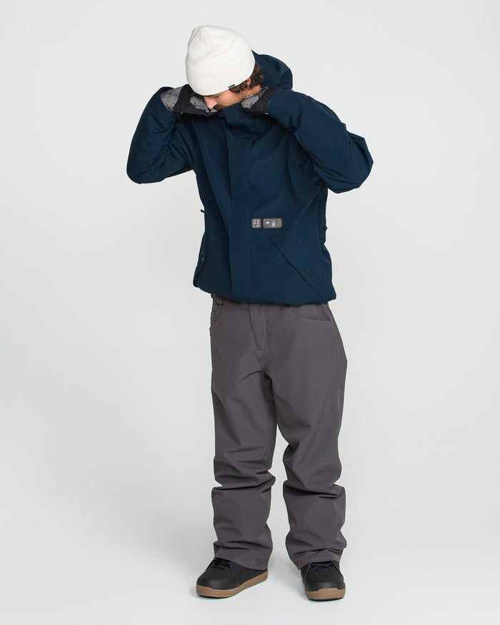 A person wearing a white beanie, gray pants, and the Volcom Men’s Double Insulated Jacket in dark blue stands against a plain white background, looking down while adjusting the hood of the jacket, which is designed for recycled warmth and features Zip Tech functionality.
