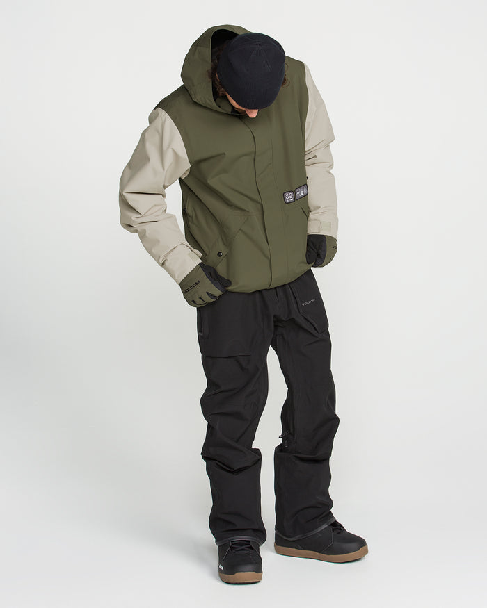 A person wearing the Volcom Men’s Double Insulated Jacket in green and beige, featuring recycled warmth, stands on a white background with hands in pockets, paired with black pants, black gloves, black shoes, and a black cap while looking down.