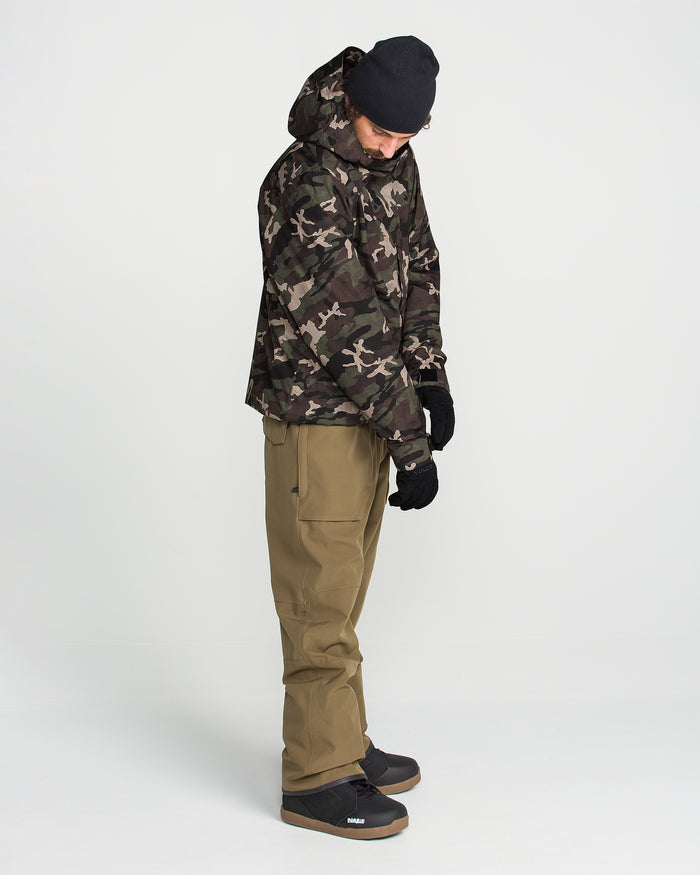 A person wearing the Volcom Men’s Double Insulated Jacket with 10K/10K V-Science technology, tan pants, black gloves, black boots, and a black beanie stands looking down against a plain white background.