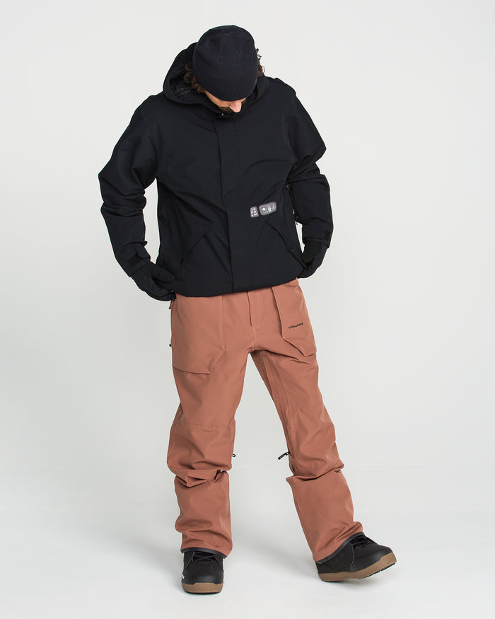 Wearing the Volcom Men’s Double Insulated Jacket, along with brown pants, black gloves, a black beanie, and black shoes, a person stands against a plain white background with hands in pockets and eyes cast downward.
