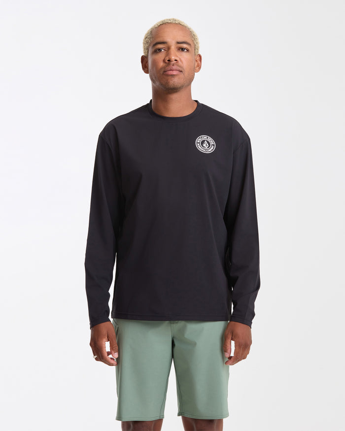 A person stands against a plain white background wearing the Volcom Men's Drain You Long Sleeve, which is black with a small white circular logo on the chest, paired with light green shorts.