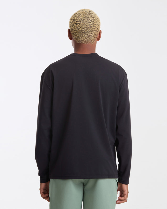 A person with short, curly blond hair is shown from the back, wearing a Volcom Men's Drain You Long Sleeve and light green pants, standing against a plain white background.