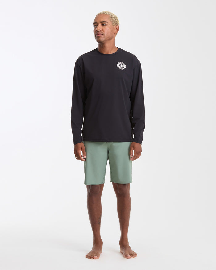 A man with short blond hair stands barefoot against a plain white background, wearing the Volcom Men's Drain You Long Sleeve black rashguard with UV protection and a small white chest logo, paired with light green shorts.