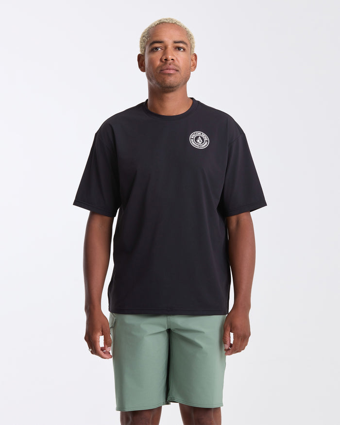 A man with short blond hair wears the Volcom Men's Drain You Short Sleeve Tee—black with a small white circular logo on the left chest—and light green shorts, posing against a plain white background.