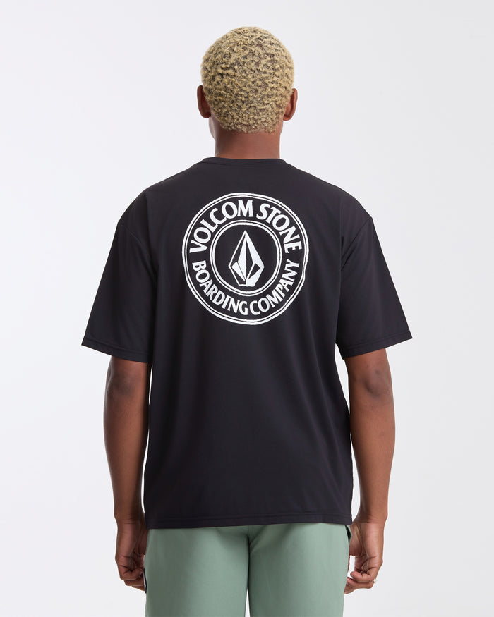 A person with short, light hair is seen from behind wearing a black Volcom Men's Drain You Short Sleeve Tee with a large white logo on the back and light green shorts. Great for UV protection during aquatic adventures. The background is plain white.