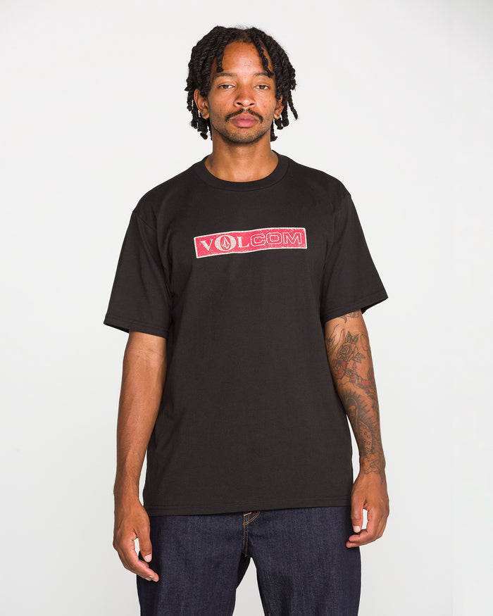 A man with tattoos stands facing the camera, wearing the Volcom Men's Dual Euro Short Sleeve Tee—a classic fit made from premium ring-spun cotton with a bold red and white vintage Volcom logo—paired with dark jeans against a plain white background.
