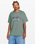 A man stands facing forward against a white background wearing the Volcom Men's Duster Short Sleeve Tee in green with a large white and black screen print, paired with light blue denim shorts. The tee is made from heavyweight organic cotton and has a loose fit.