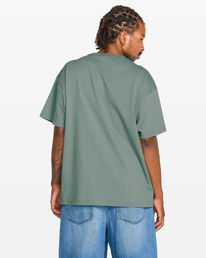 A man with braided hair, seen from behind, wears the Volcom Men's Duster Short Sleeve Tee in green—crafted from heavyweight organic cotton—paired with light blue denim shorts against a plain light background.