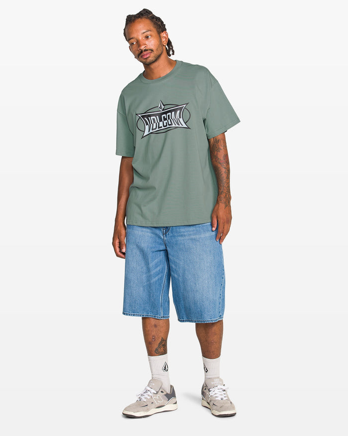 A man with tattooed arms stands against a plain background wearing the Volcom Men's Duster Short Sleeve Tee, loose denim shorts, white socks, and gray sneakers, looking slightly to the side.