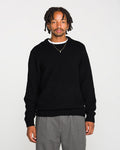 A man with short dreadlocks wears the Volcom Men's Edmonder II Sweater in black, paired with gray pants and a gold necklace. He stands facing forward against a plain white background.