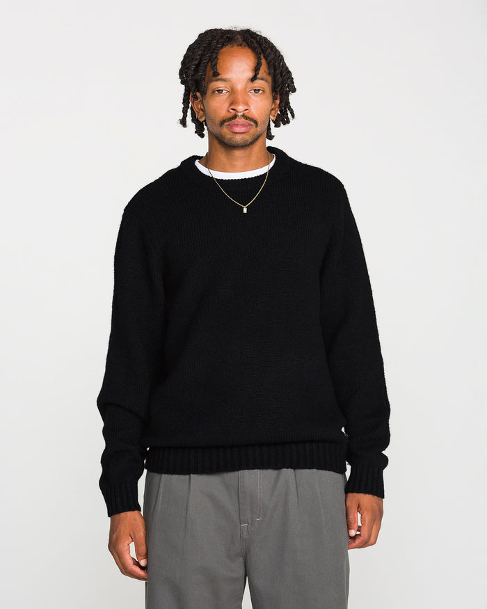 A man with short dreadlocks wears the Volcom Men's Edmonder II Sweater in black, paired with gray pants and a gold necklace. He stands facing forward against a plain white background.