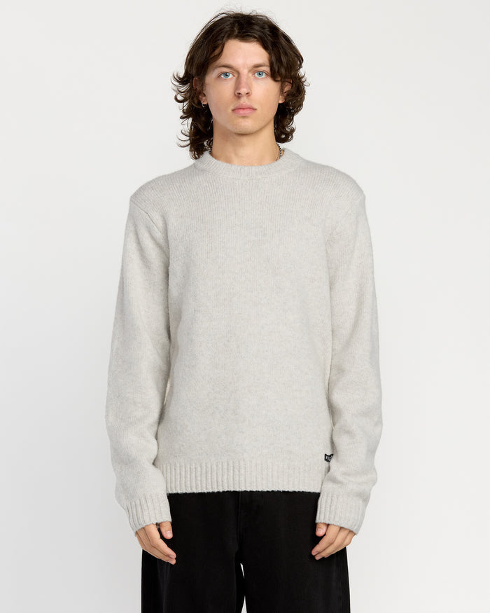 A man with wavy brown hair wears the Volcom Men's Edmonder II Sweater in light gray and loose black pants, standing facing forward against a plain white background.