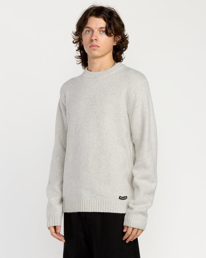A person with shoulder-length wavy hair wears the Volcom Men's Edmonder II Sweater in light gray and black pants, standing against a plain white background.