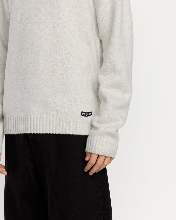 A person wears Volcom's Men's Edmonder II Sweater in light gray, featuring a small black brand tag near the hem, paired with loose black pants. Only the lower torso and one hand appear against a plain white background.
