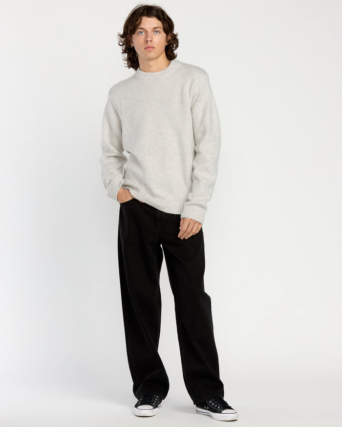 A person with medium-length brown hair wears the Volcom Men's Edmonder II Sweater in light gray, paired with loose black pants and black sneakers, standing against a plain white background with one hand in their pocket.