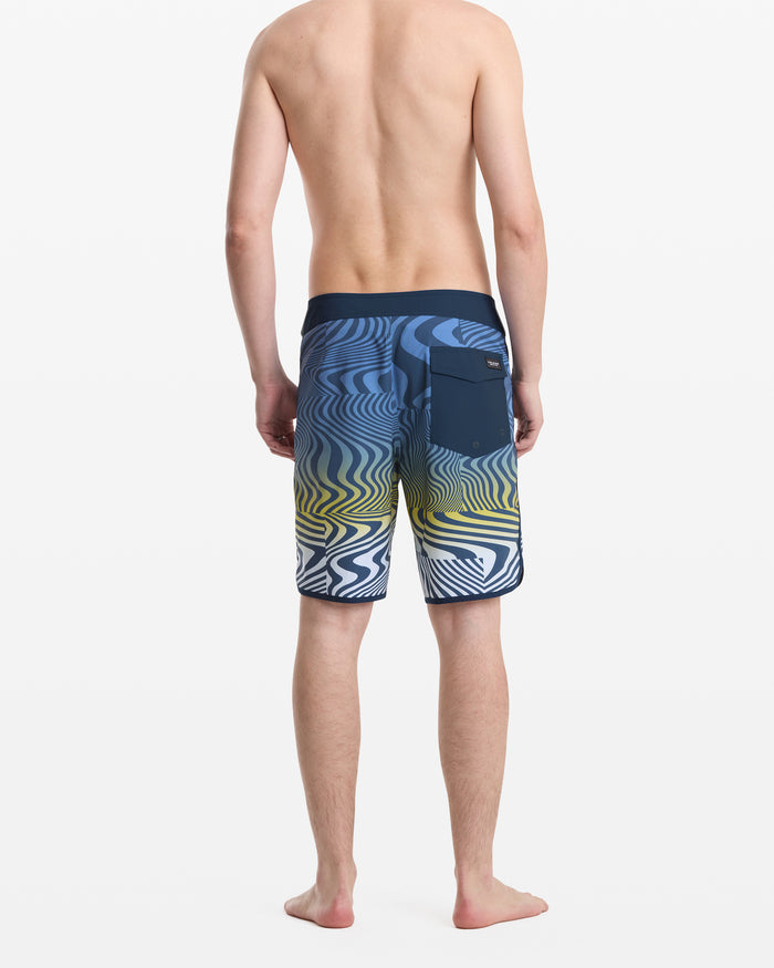 A barefoot man faces away, wearing Volcom Men's Encounter Scallop Mod 19 board shorts featuring a blue, yellow, and white wavy design with a right back pocket. He is shirtless against a plain white background.
