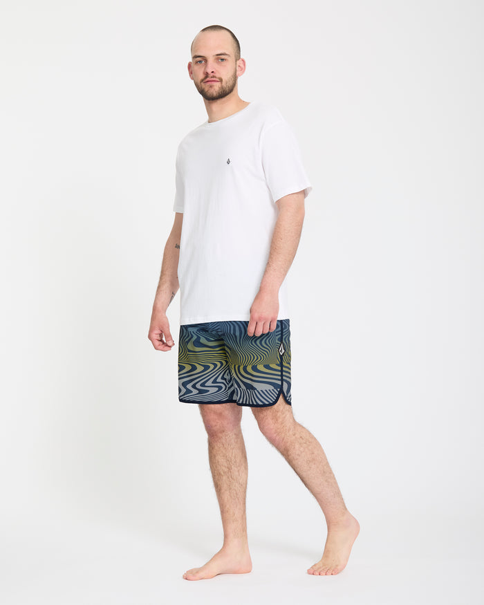 A barefoot man with a beard stands against a plain white background, wearing a white t-shirt and Volcom Men's Encounter Scallop Mod 19 shorts featuring blue, green, and yellow swirling patterns.