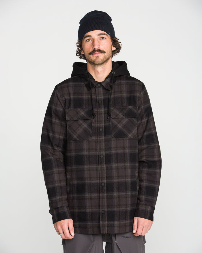 A man with a mustache, wearing a black beanie, Volcom Men’s Enson Insulated Riding Flannel in a black and gray plaid pattern, and gray pants stands facing forward against a plain white background.