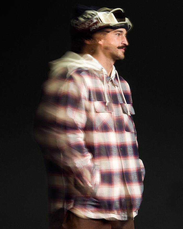 A man stands against a dark background, wearing the Volcom Men’s Enson Insulated Riding Flannel over a white hoodie with a fleece hood and ski goggles. A blur effect gives his left side a sense of motion.