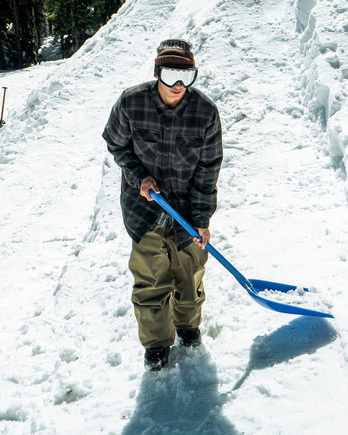 Wearing the Volcom Men’s Enson Insulated Riding Flannel, a person dressed in winter attire uses a blue snow shovel to move snow outdoors on a sunny day.