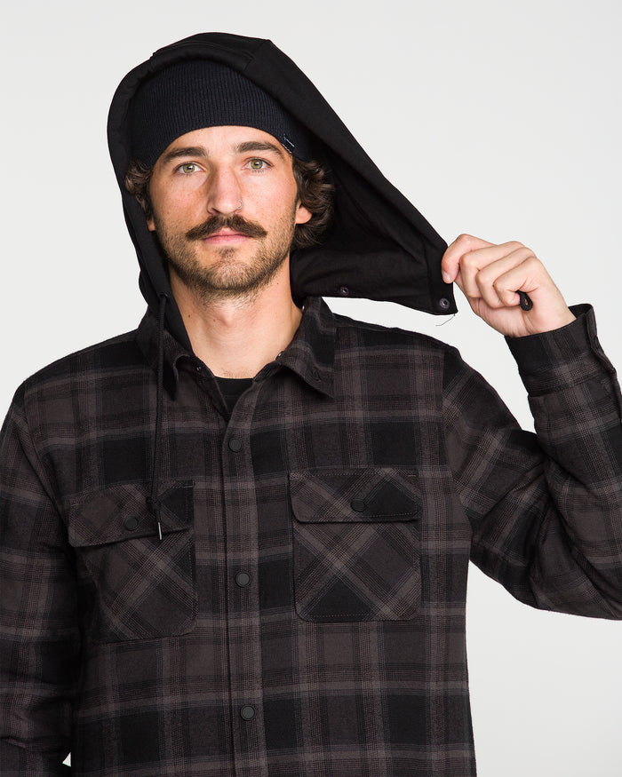 A man with a mustache wearing a black beanie and the Volcom Men’s Enson Insulated Riding Flannel holds up the fleece hood of his jacket with one hand, looking directly at the camera against a plain background.