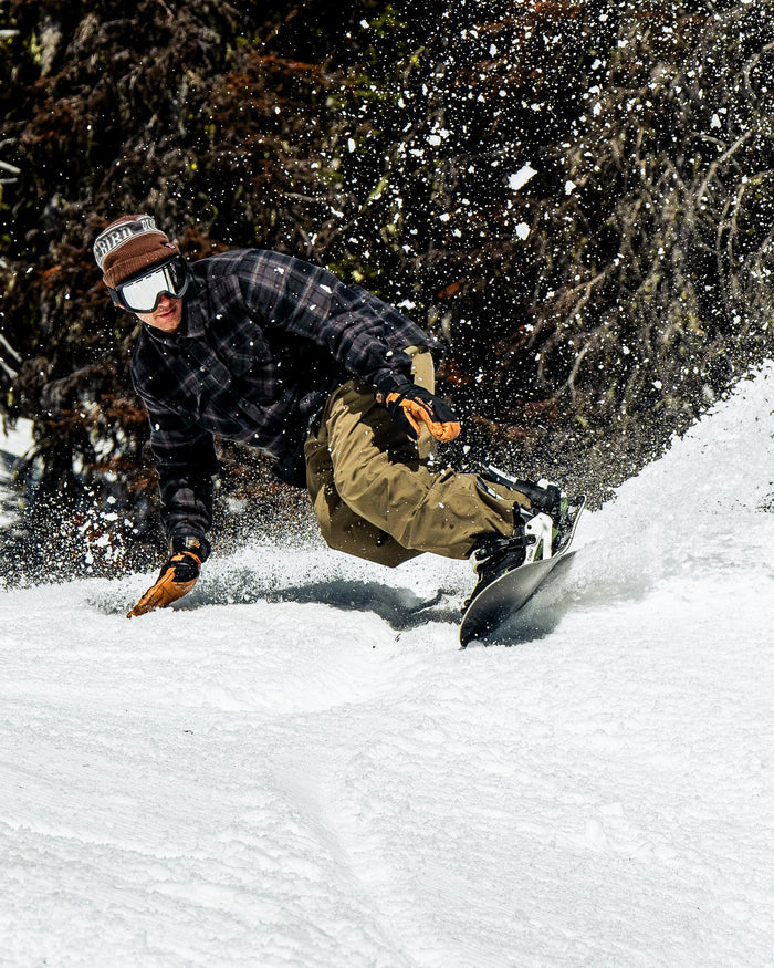 Wearing the Volcom Men’s Enson Insulated Riding Flannel, a snowboarder—also sporting goggles and a hat over their winter gear—carves a sharp turn down a snowy slope, kicking up powder near the trees.