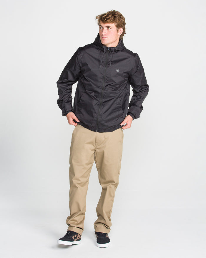 A man wearing the Volcom Men's Ermont Jacket in black, paired with beige pants and black sneakers, stands against a plain white background, looking to his left with his hands near the jacket pockets.