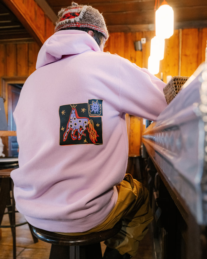 A person wearing the Volcom Men’s Essential Hoodie, featuring a pink cotton-poly fleece and a colorful giraffe patch, sits at a wooden bar facing away. The setting is warm and cozy, illuminated by yellow lighting with wooden walls.