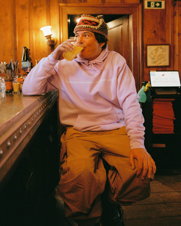 A person wearing a colorful knit hat, tan pants, and a Volcom Men’s Essential Hoodie in light purple cotton-poly fleece sits at a bar, sipping a drink from a glass, with wood-paneled walls and bottles lining the shelves in the background—a scene reminiscent of Arthur Longo’s relaxed style.
