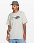 A man stands against a plain white background wearing a light gray Volcom Men's Eurobox Short Sleeve Tee and a gray Volcom cap. He has tattoos on his right arm, blue jeans, and looks slightly to the side.