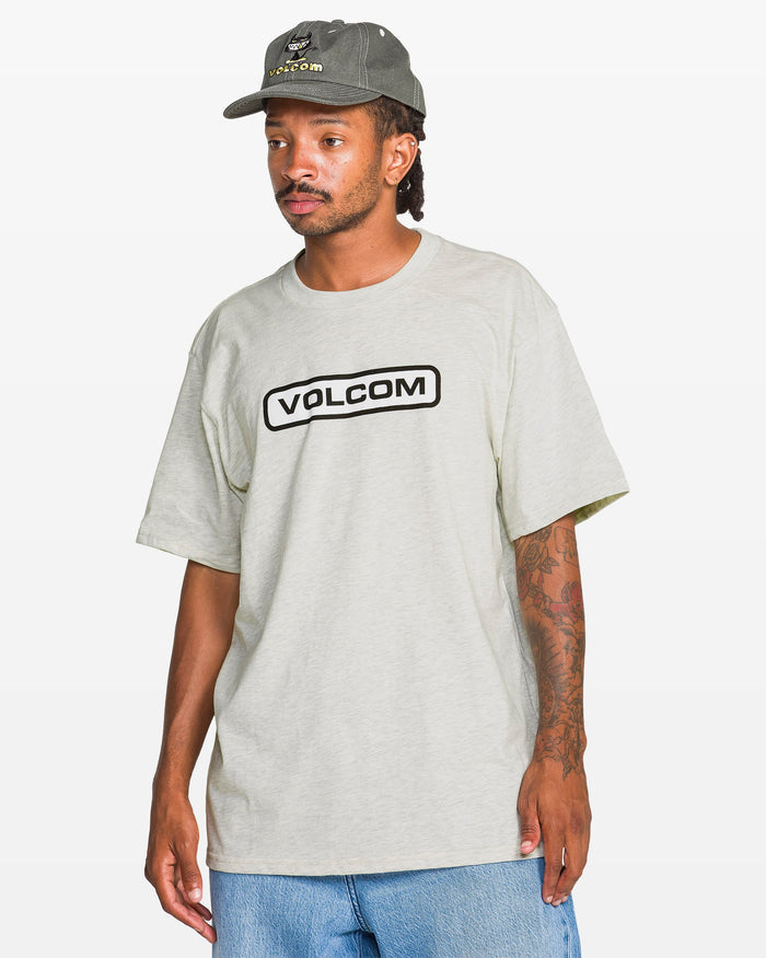 A man stands against a plain white background wearing a light gray Volcom Men's Eurobox Short Sleeve Tee and a gray Volcom cap. He has tattoos on his right arm, blue jeans, and looks slightly to the side.