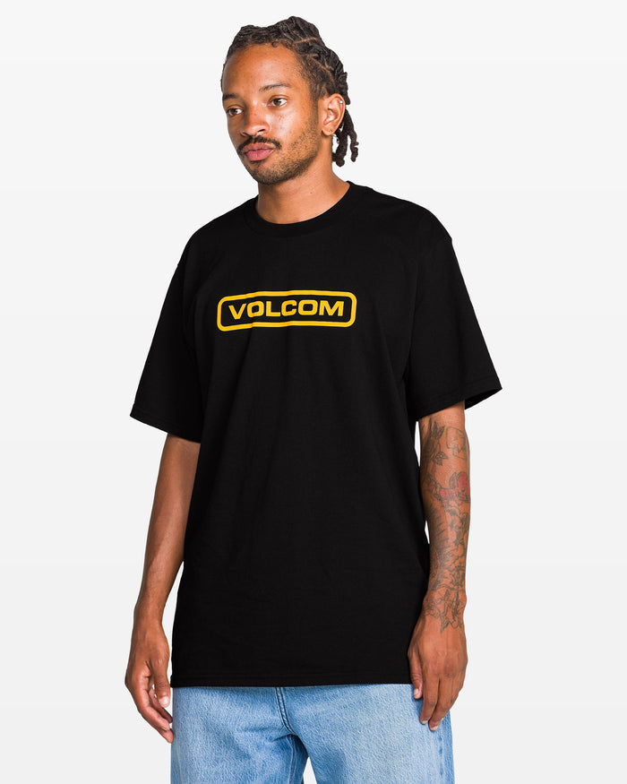 A man with braided hair and tattoos on his left arm wears the Volcom Men's Eurobox Short Sleeve Tee, a loose 100% cotton black crew neck shirt with a yellow logo, paired with light blue jeans, standing against a plain white background.