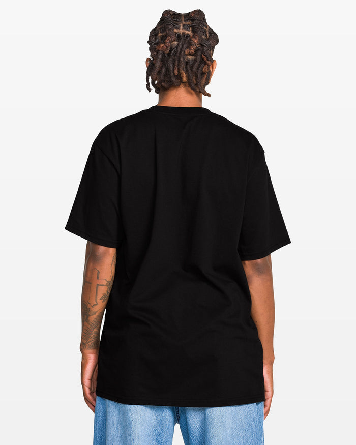 Person with short dreadlocks, seen from the back in a plain black Volcom Men's Eurobox Short Sleeve Tee (100% cotton) and light blue jeans, against a white background. Tattoo is visible on left arm.