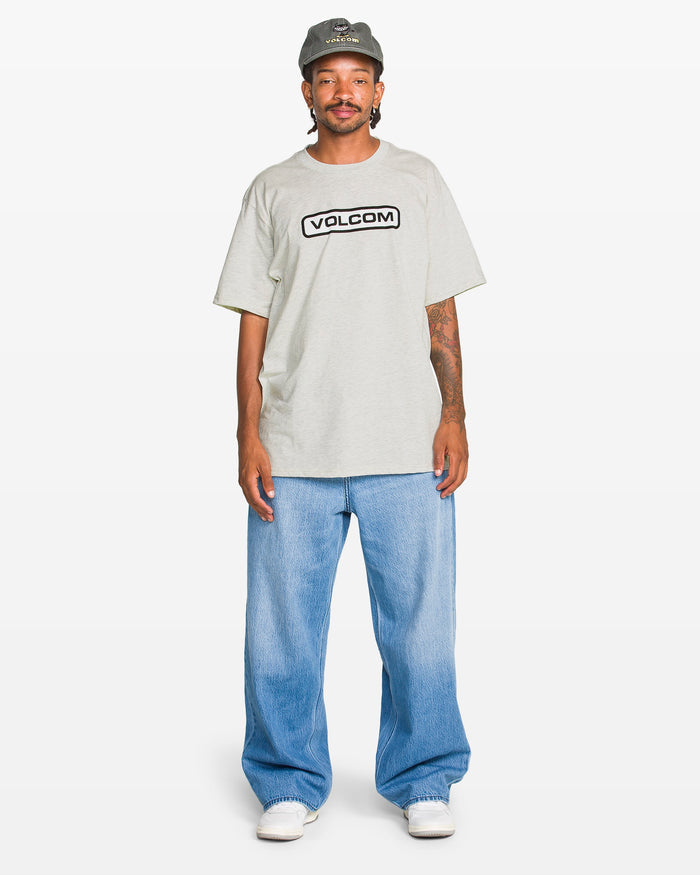 A person wears a gray Volcom Men's Eurobox Short Sleeve Tee with a soft-hand print, light blue baggy jeans, white sneakers, and a gray cap while standing against a plain white background.
