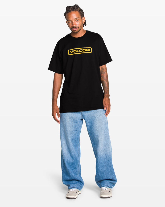 A man faces forward wearing the Volcom Men's Eurobox Short Sleeve Tee in black with a yellow logo, light blue baggy jeans, and white sneakers against a plain white background.